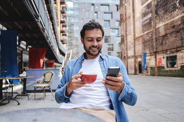 Lächelnder Mann, der eine Kaffeetasse hält und sein Smartphone in einem Straßencafé benutzt - ASGF04664