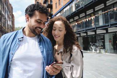 Smiling woman with man using smart phone at street on sunny day - ASGF04666