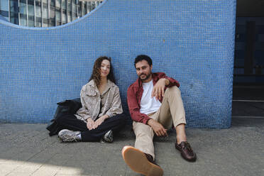 Lächelnder Mann und lächelnde Frau sitzen auf dem Boden in der Nähe eines Gebäudes - ASGF04677