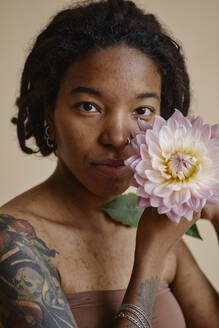 Young woman with tattoo and acne scars holding flower against brown background - KPEF00262