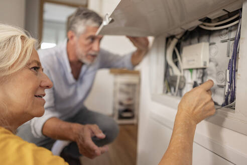 Woman examining electrical cable in fuse box with man at home - JCCMF10744