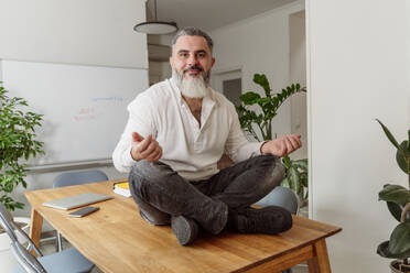 Smiling businessman meditating in lotus position on table at office - OSF02182