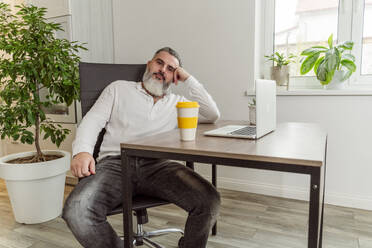 Thoughtful businessman with reusable coffee and laptop on table at work place - OSF02183