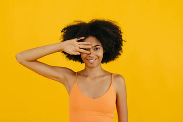 Smiling woman covering eye with hand against yellow background - TCEF02285