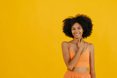 Smiling woman touching neck standing against yellow background - TCEF02286