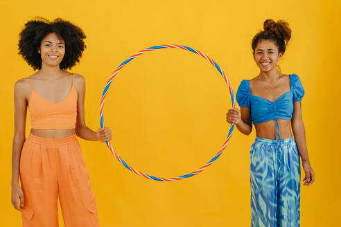 Smiling multiracial friends holding plastic hoop standing against yellow background - TCEF02313