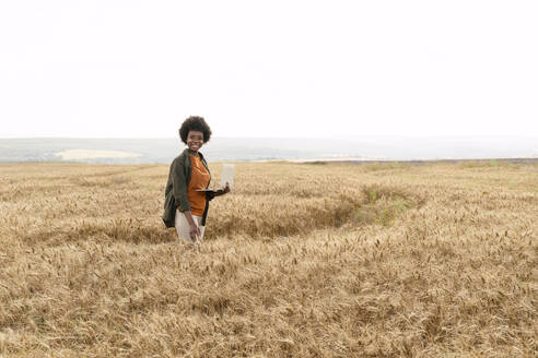Smiling agronomist holding laptop standing amidst barley crop at field - AAZF01153