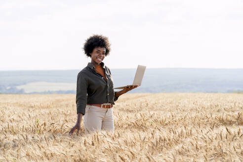 Lächelnder Afro-Agronom mit Laptop inmitten von Gerstenkulturen auf dem Feld - AAZF01158