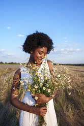 Happy young woman holding bunch of daises in field - AAZF01209