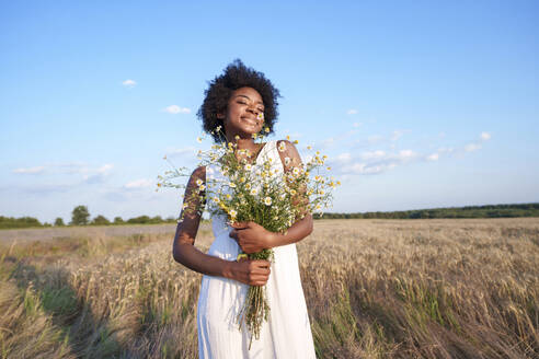 Smiling young woman holding bunch of daises in field under sky - AAZF01210