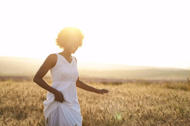 Fröhliche junge Frau läuft in einem Weizenfeld bei Sonnenuntergang - AAZF01216