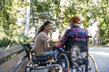 Smiling caregiver consoling retired senior man in wheelchair at park - IKF01320