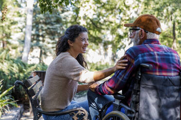 Cheerful caregiver consoling retired senior man in wheelchair at park - IKF01321