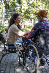 Smiling caregiver sitting on bench by retired senior man in wheelchair at park - IKF01322