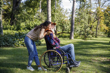 Happy caregiver embracing retired senior man in wheelchair at park - IKF01331