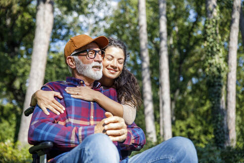 Smiling caregiver embracing retired senior man in wheelchair at park - IKF01335