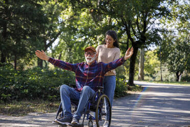 Social worker pushing cheerful retired senior man with arms outstretched in wheelchair on road at park - IKF01341