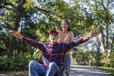 Volunteer pushing cheerful retired senior man with arms outstretched in wheelchair on road at park - IKF01342