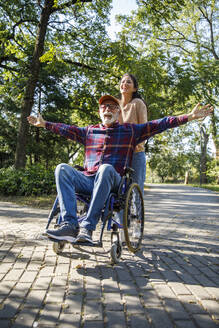 Caregiver pushing cheerful retired senior man with arms outstretched in wheelchair on road at park - IKF01343