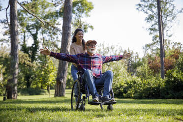 Social worker pushing cheerful retired senior man with arms outstretched in wheelchair on grass at park - IKF01344