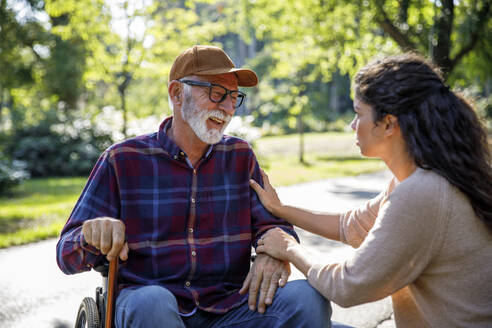 Happy retired senior man in wheelchair talking with social worker at park - IKF01347