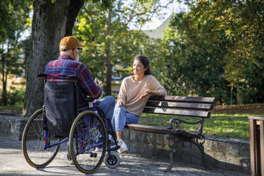 Smiling caregiver sitting on bench listening to retired man in wheelchair at park - IKF01349