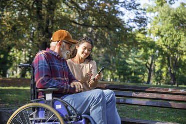 Smiling caregiver showing smart phone with senior man in wheelchair at park - IKF01350