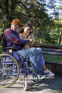 Smiling caregiver sharing smart phone with senior man in wheelchair at park - IKF01351