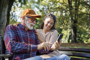 Senior man in wheelchair at park showing smiling caregiver showing smart phone - IKF01352