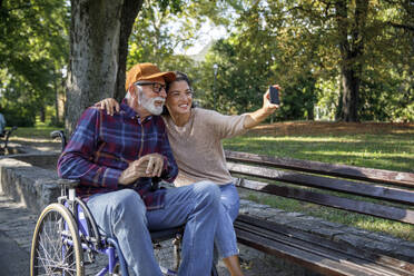 Smiling caregiver taking selfie with senior man in wheelchair at park - IKF01353