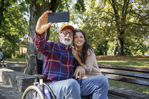 Smiling senior man in wheelchair taking selfie with social worker at park - IKF01357