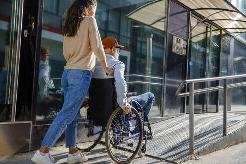 Caregiver pushing senior man in wheelchair on ramp - IKF01361