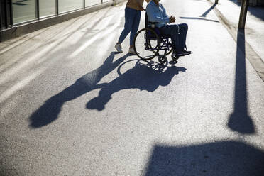 Caregiver pushing man in wheelchair on footpath with shadow - IKF01362