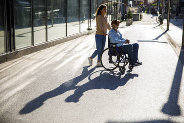 Volunteer pushing man in wheelchair on footpath with shadow - IKF01363