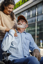 Smiling caregiver with hand on shoulder looking at senior man in wheelchair - IKF01368