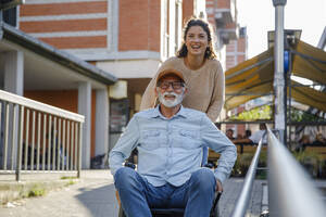 Happy caregiver pushing retired man in wheelchair by railing - IKF01370