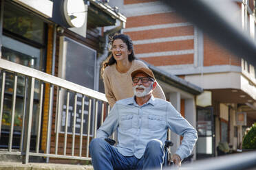 Happy volunteer pushing retired man in wheelchair by railing - IKF01371