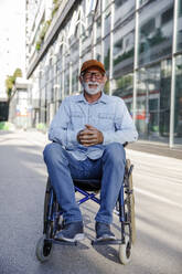 Retired senior man with hands clasped sitting in wheelchair on footpath - IKF01372