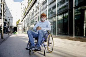 Smiling retired senior man with hands clasped sitting in wheelchair on footpath - IKF01373