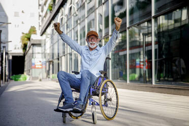 Happy senior man with arms raised sitting in wheelchair - IKF01376