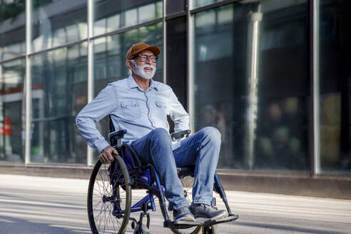 Smiling retired senior man sitting in wheelchair on footpath - IKF01381