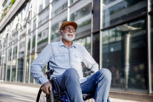 Smiling senior man sitting in wheelchair on footpath - IKF01382