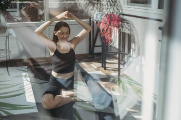 Frau mit erhobenen Armen macht Yoga auf Matte zu Hause durch Glas gesehen - ANAF02174