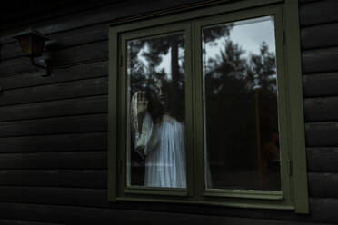 Female ghost looking through glass window of haunted cabin - MASF39569