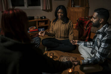 Gemischtrassige Freunde halten sich an den Händen und sitzen mit einem Ouija-Brett in einer Blockhütte - MASF39581