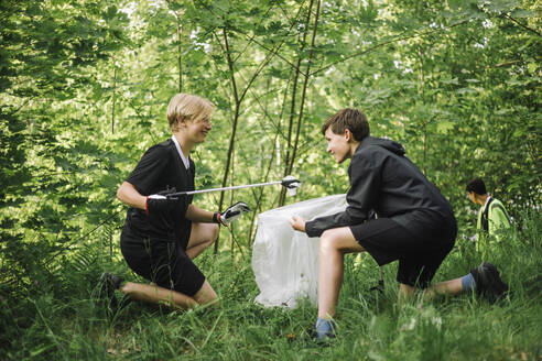 Full length of smiling boy kneeling while collecting plastic together - MASF39670