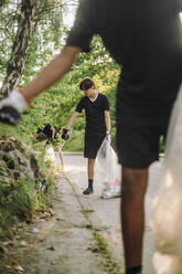 Teenage boys collecting plastic from plants in garbage bags on road - MASF39710