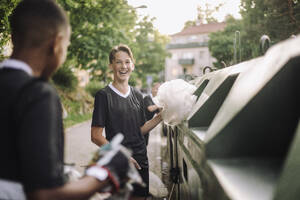 Glücklicher Teenager, der einen Plastikmüllsack in eine Recycling-Tonne in der Nähe von Freunden steckt - MASF39726