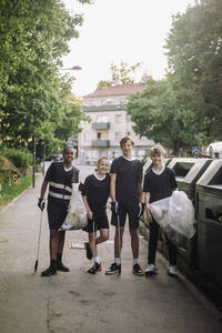 Ganzkörperporträt von lächelnden Jungen, die mit Müllsäcken neben Recycling-Tonnen stehen - MASF39729