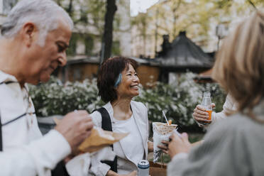 Smiling senior woman enjoying snacks with friends while standing at street - MASF39747